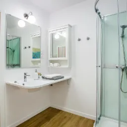 Jasna łazienka z drewnianymi elementami: drzwi z naturalnego drewna, podłoga w panelach imitujących drewno, biała umywalka z szafką i lustrem, kabina prysznicowa z mlecznego szkła.