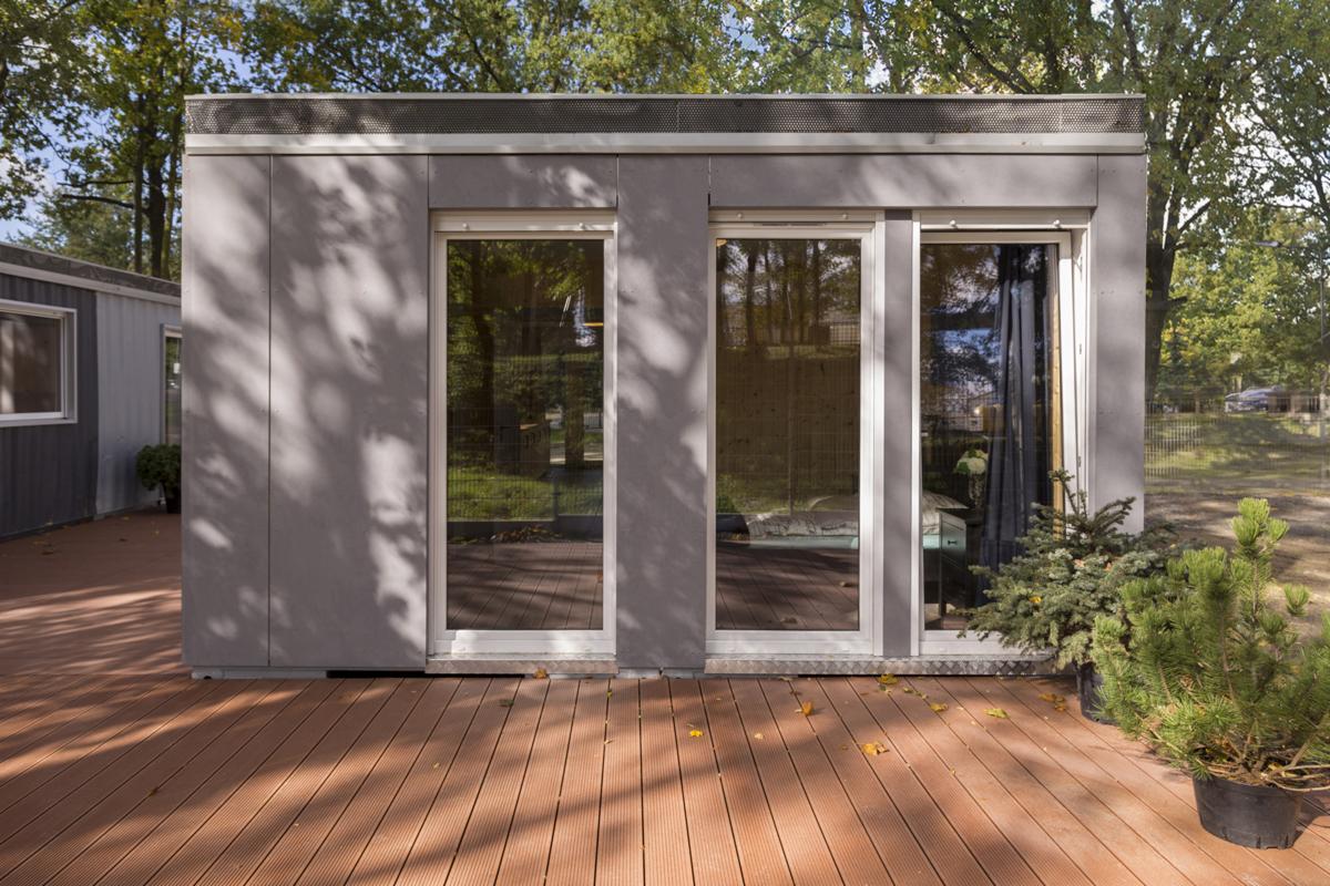 Nowoczesny, modułowy budynek z szarej elewacji z trzema wysokimi oknami, usytuowany na drewnianym tarasie, otoczony zielenią drzew.