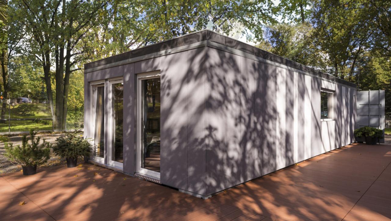 Nowoczesny, parterowy budynek o szarej elewacji z dużymi oknami, otoczony zielenią drzew i drewnianym tarasem. Na tarasie widoczne małe drzewka iglaste w czarnych donicach.