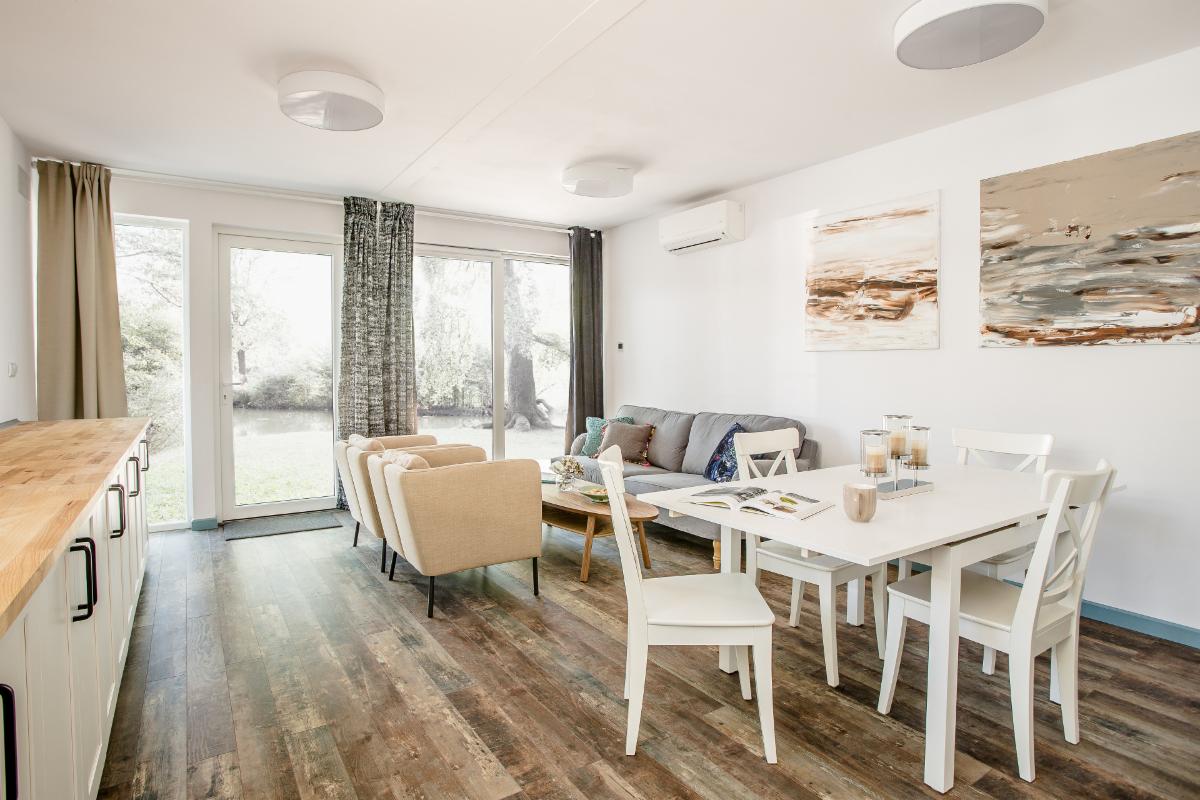 Jasne wnętrze domu z drewnopodobną podłogą, beżowymi fotelami i białym stołem jadalnym, widok na ogród przez duże okna.