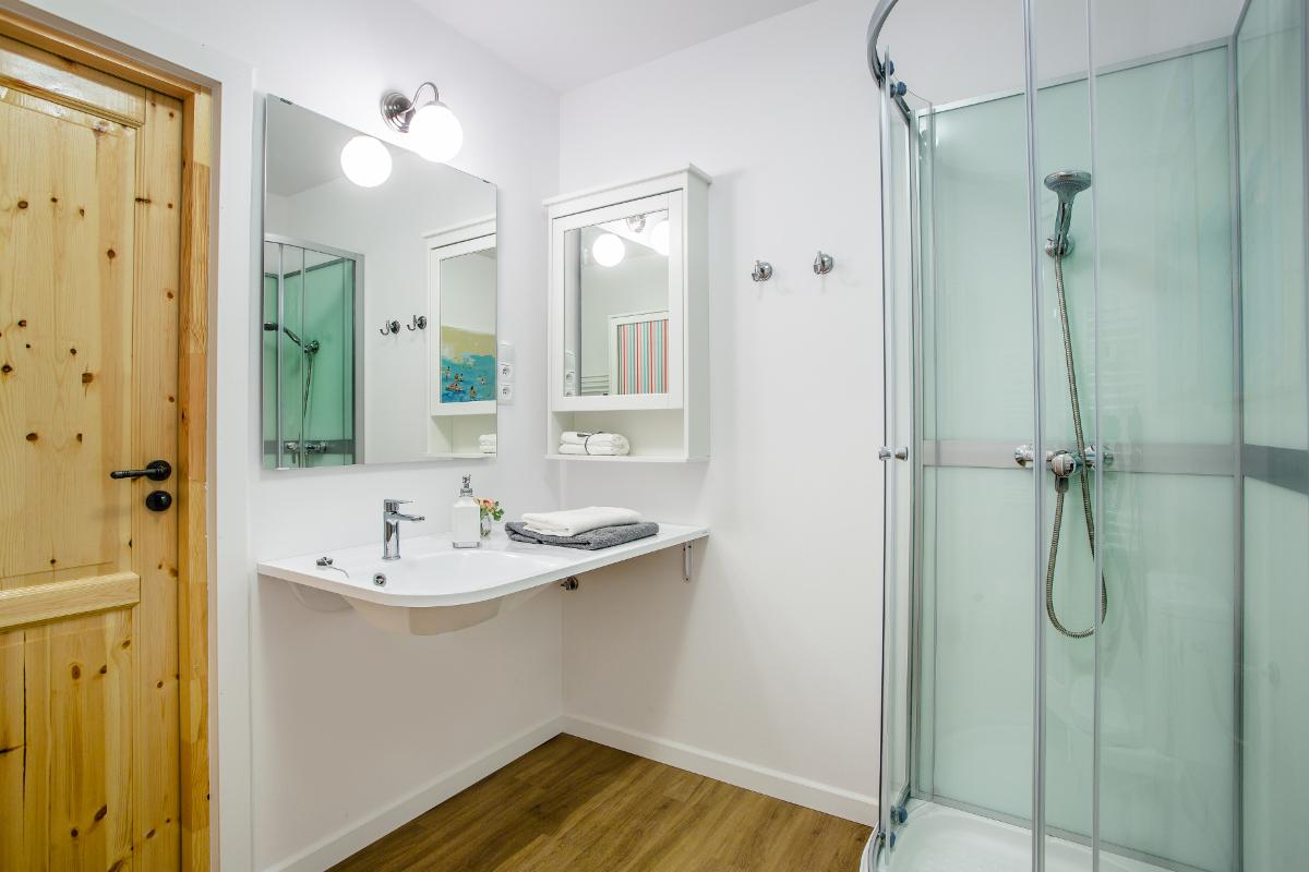 Jasna łazienka z drewnianymi elementami: drzwi z naturalnego drewna, podłoga w panelach imitujących drewno, biała umywalka z szafką i lustrem, kabina prysznicowa z mlecznego szkła.