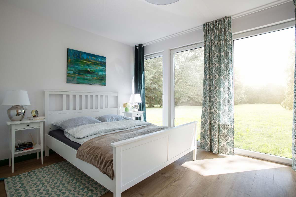 Jasne wnętrze sypialni w domu szkieletowym z białym drewnianym łóżkiem, szafką nocną i widokiem na ogród przez duże okna z wzorzystymi zasłonami.