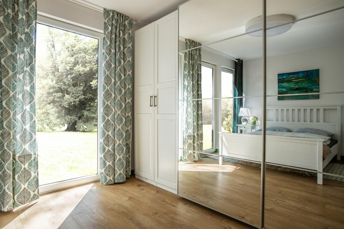 Jasne wnętrze sypialni z drewnianą podłogą, białym łóżkiem i szafą z lustrzanymi drzwiami, okno z zasłonami w liście.