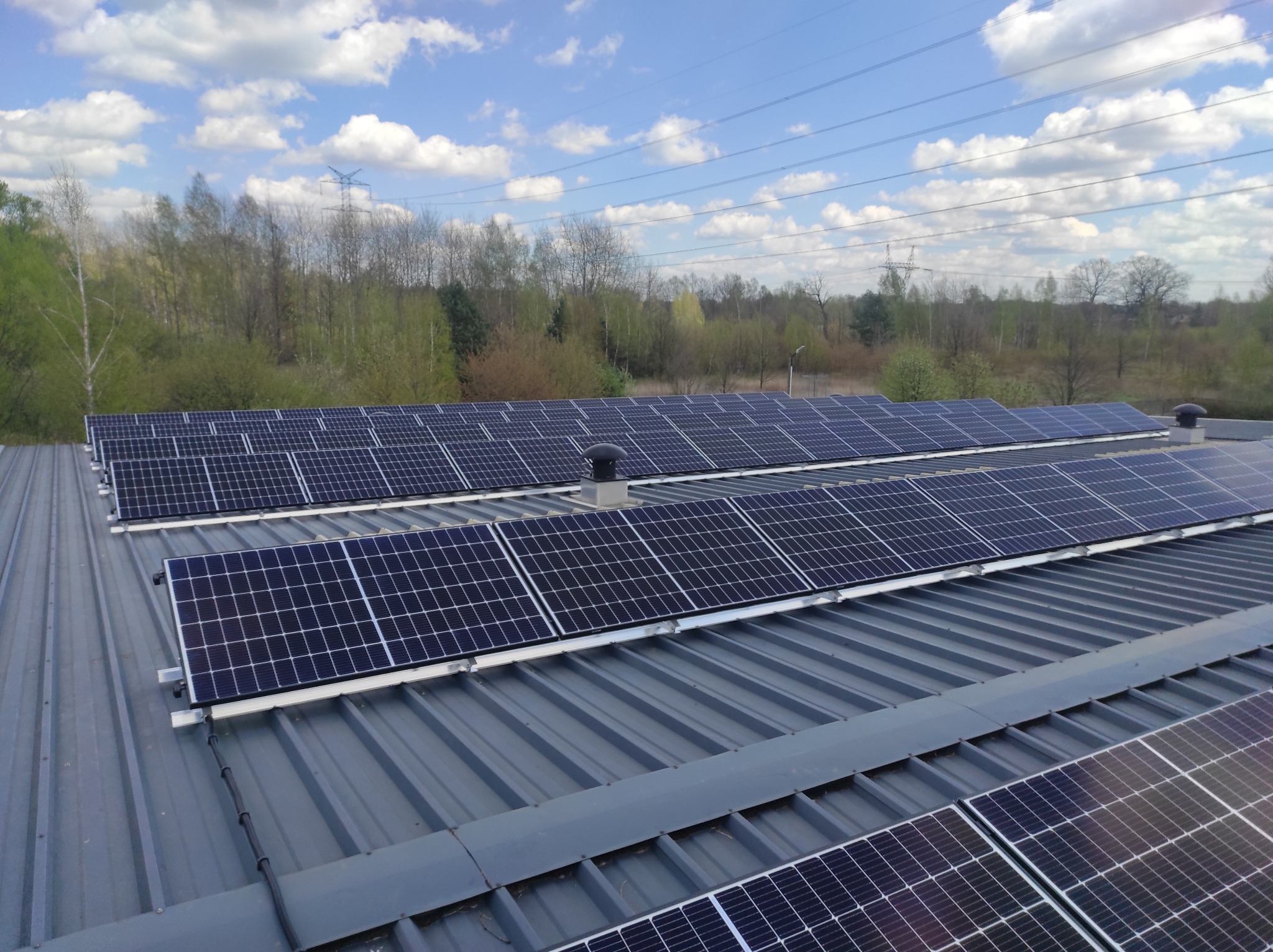 Rząd paneli słonecznych na blaszanym dachu budynku, z kominkiem wentylacyjnym i liniami wysokiego napięcia w tle, przy częściowo zachmurzonym niebie.