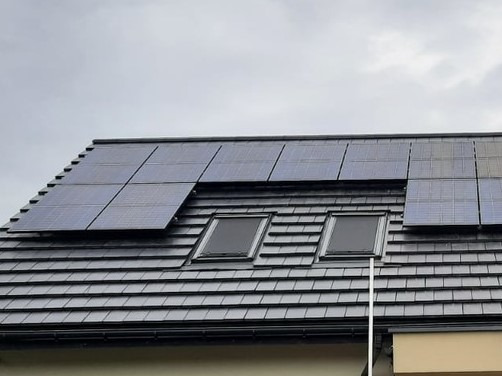 Panele słoneczne na dachu z dachówki ceramicznej, dwa okna dachowe, pochmurne niebo w tle.