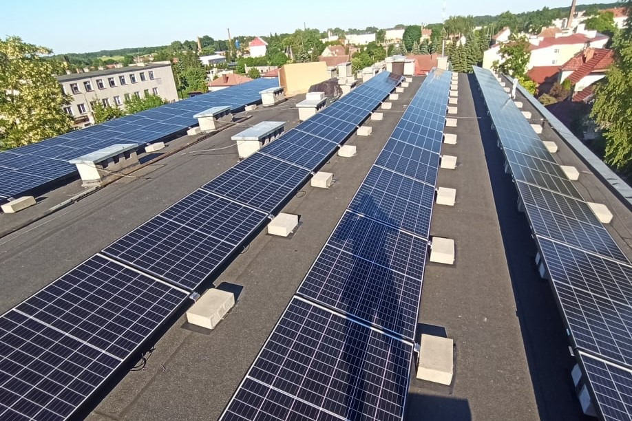 Rząd paneli fotowoltaicznych na płaskim dachu budynku przemysłowego, widok z góry, betonowe podpory, w tle zabudowa miejska i drzewa.