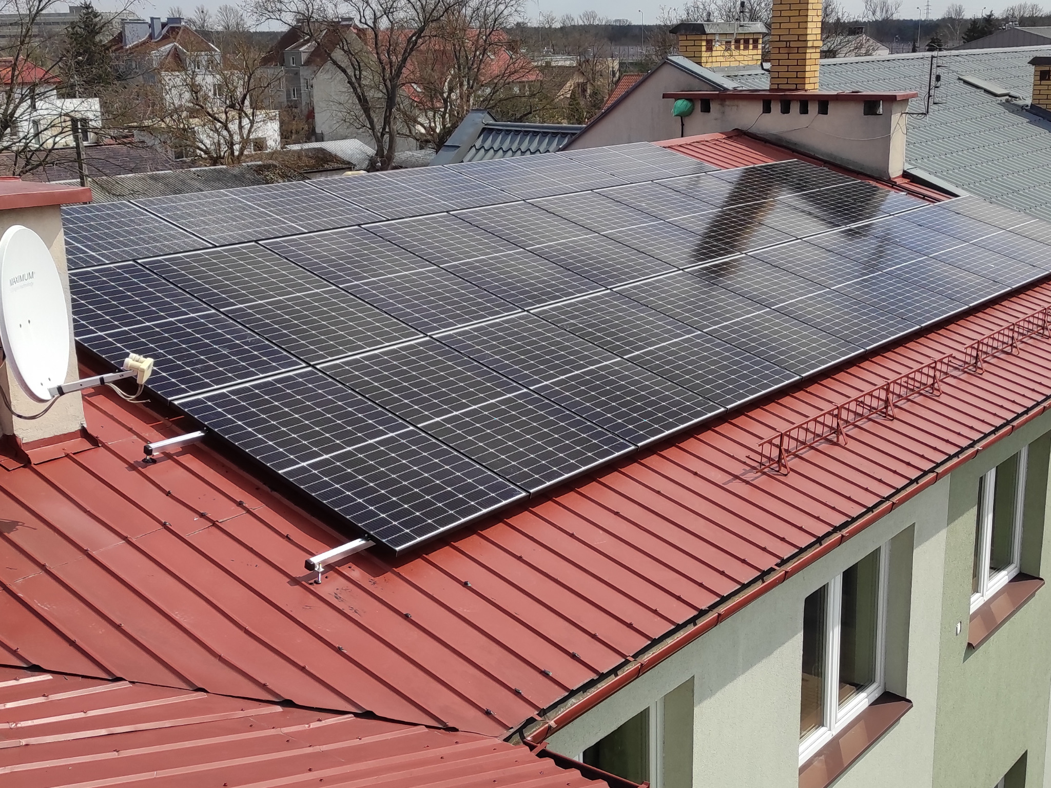 Panele słoneczne na czerwonym, blaszanym dachu budynku wielorodzinnego, z widoczną anteną satelitarną na ścianie szczytowej.