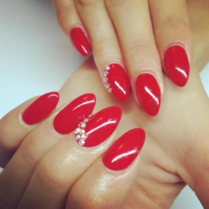 Czerwone, migdałowe paznokcie z drobnymi kryształkami ułożonymi wzdłuż boku dwóch paznokci.