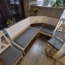 Kącik jadalny z narożną ławką z białego drewna i szarymi siedziskami, uzupełniony o krzesła w stylu skandynawskim i drewnianą podłogę imitującą deski.