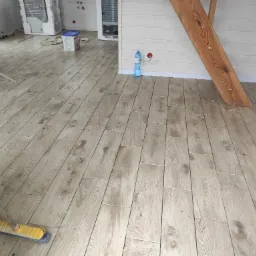 Ułożone panele podłogowe imitujące drewno w jasnym wnętrzu w Gowidlinie, widoczna szczotka do zamiatania i butelka wody mineralnej.