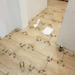 Układanie płytek podłogowych imitujących drewno z użyciem żółtych krzyżyków dystansowych, widoczne narzędzia, puszka napoju i notatki na podłodze.