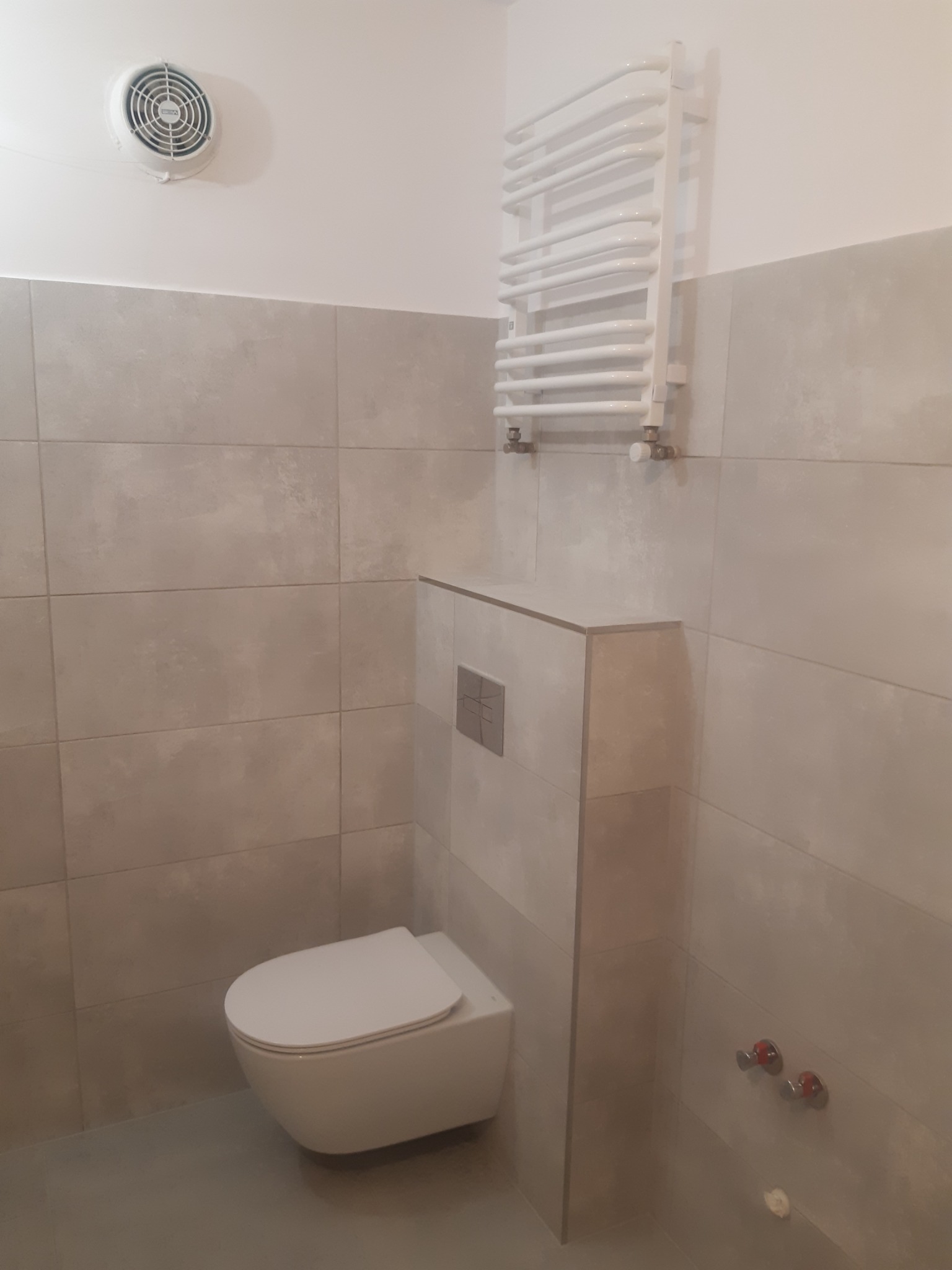 Wykończona łazienka z beżowymi płytkami, podwieszaną toaletą i grzejnikiem drabinkowym. Widoczny wentylator sufitowy i zawory. Minimalistyczny design.