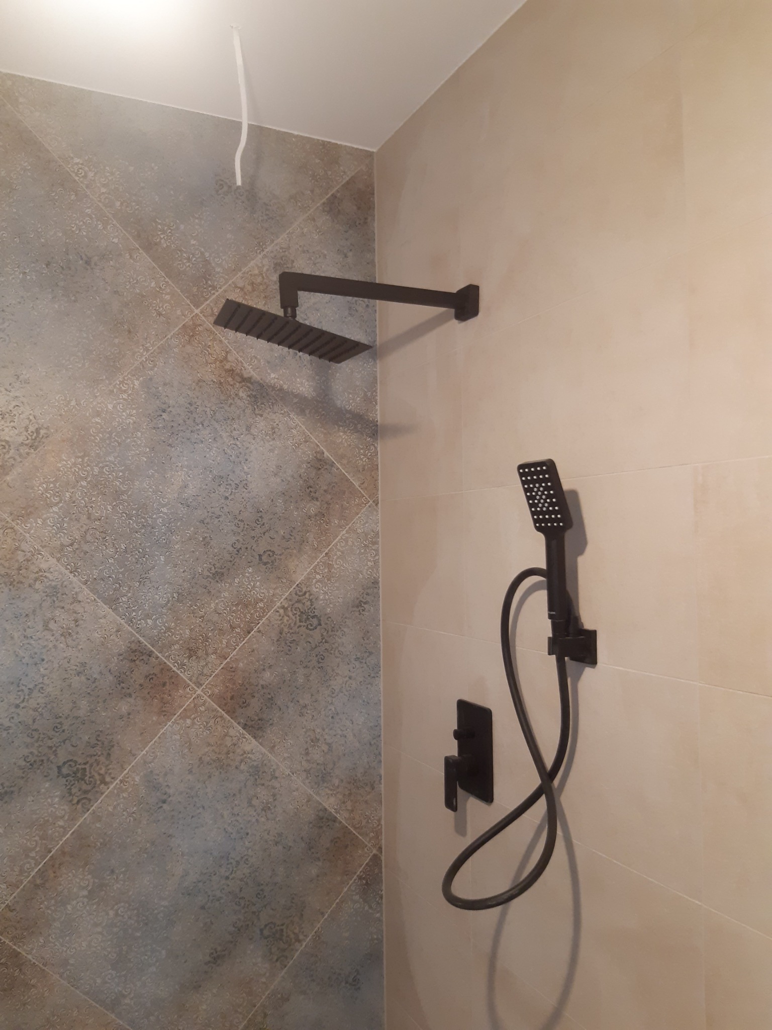 Wykończona łazienka z nowoczesną, czarną armaturą: deszczownica i słuchawka prysznicowa na tle beżowych i wzorzystych płytek. Widoczny fragment sufitu z wystającym kablem.