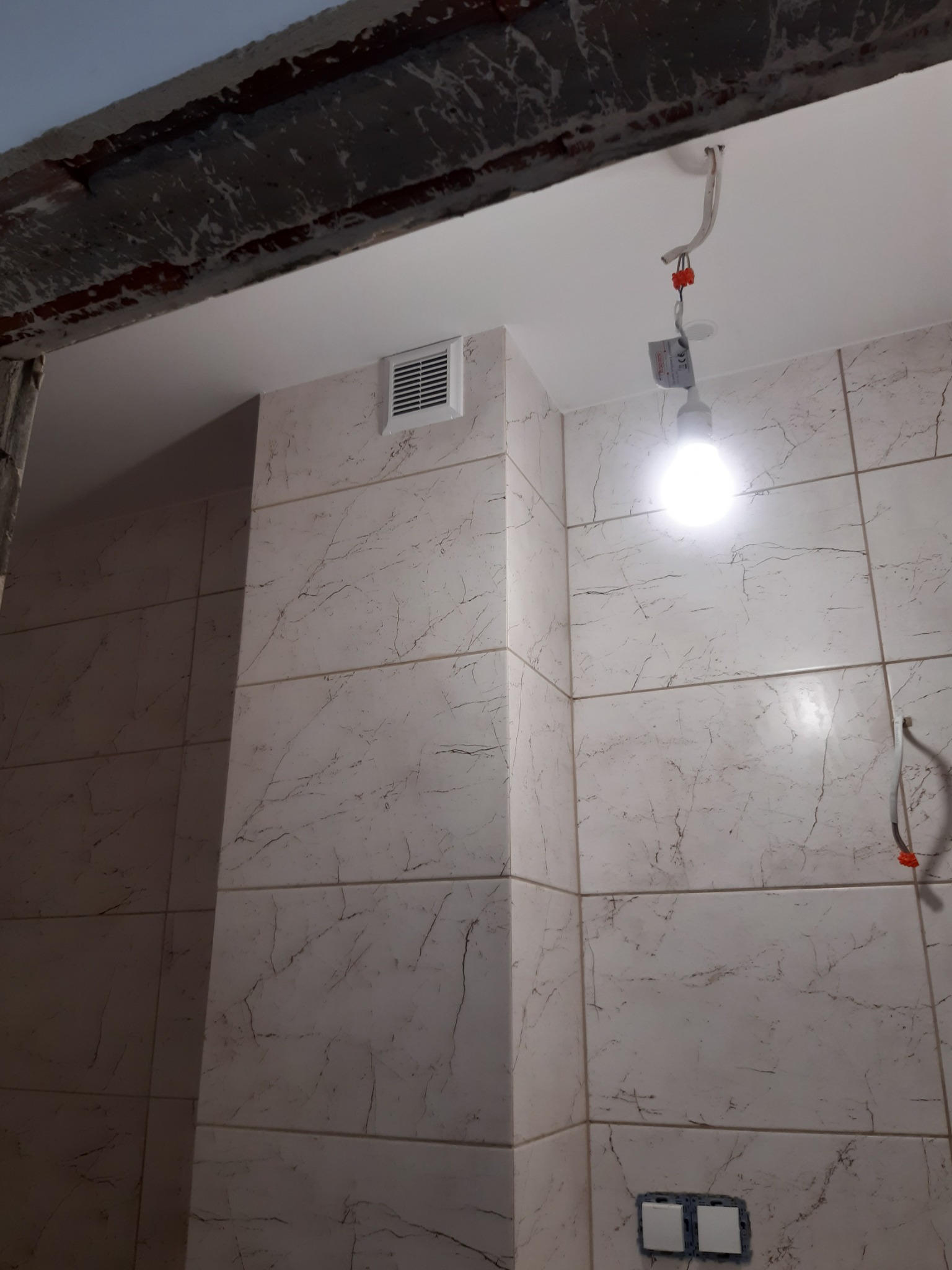 Wykończona łazienka z marmurowymi płytkami, widoczna wentylacja i instalacja elektryczna z żarówką. Fragment surowego wykończenia nad drzwiami.