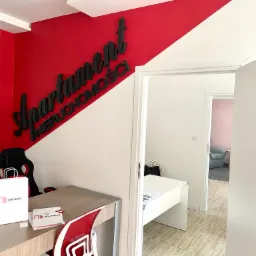 Wnętrze biura agencji nieruchomości z logo na ścianie, biurkiem z torbą reklamową i widokiem na kolejne pomieszczenie z fotelem.