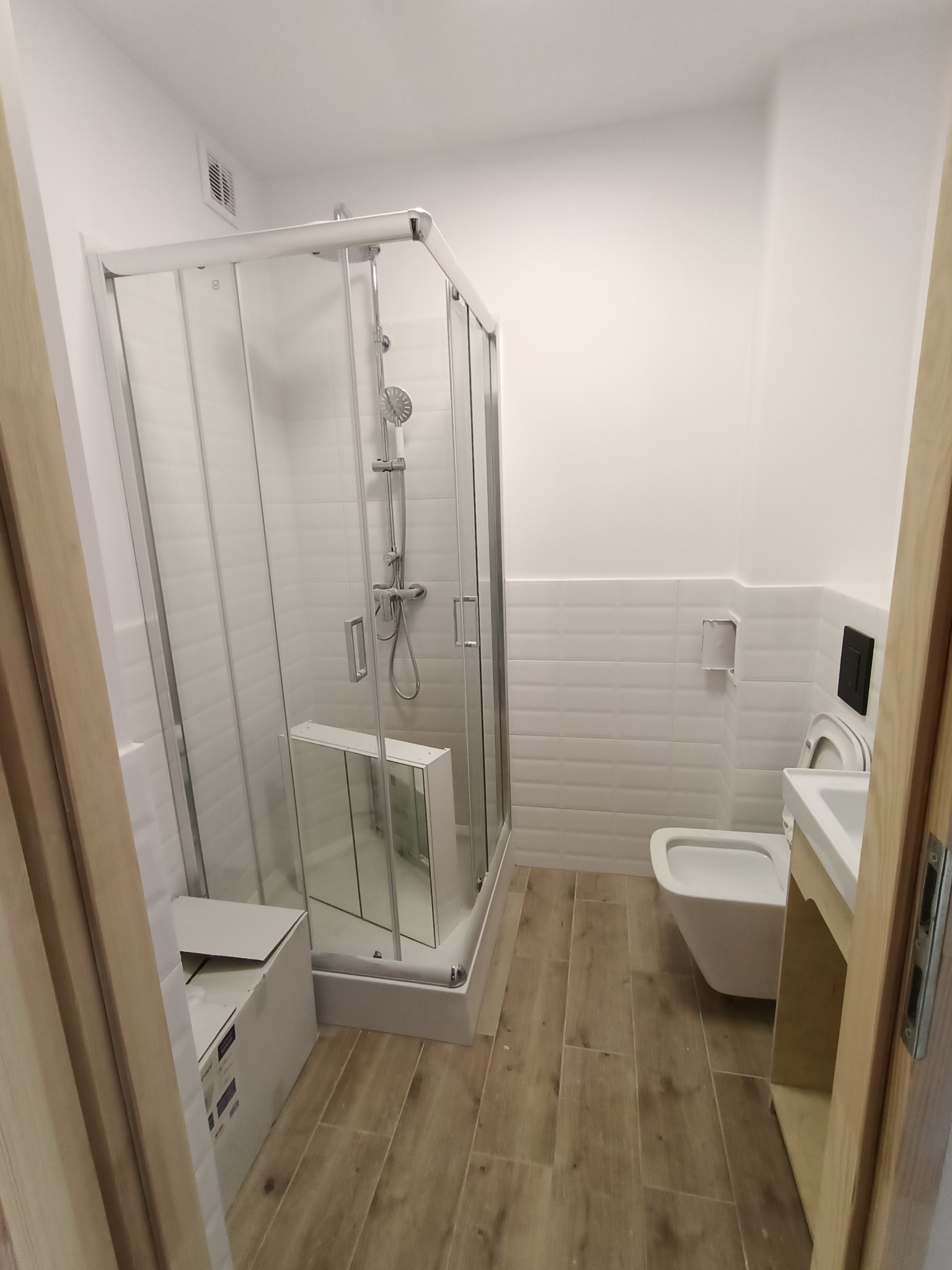 Nowoczesna łazienka z kabiną prysznicową, wiszącą toaletą i umywalką z szafką, wykończona białymi płytkami i panelami podłogowymi imitującymi drewno. W rogu widoczne kartonowe opakowanie.