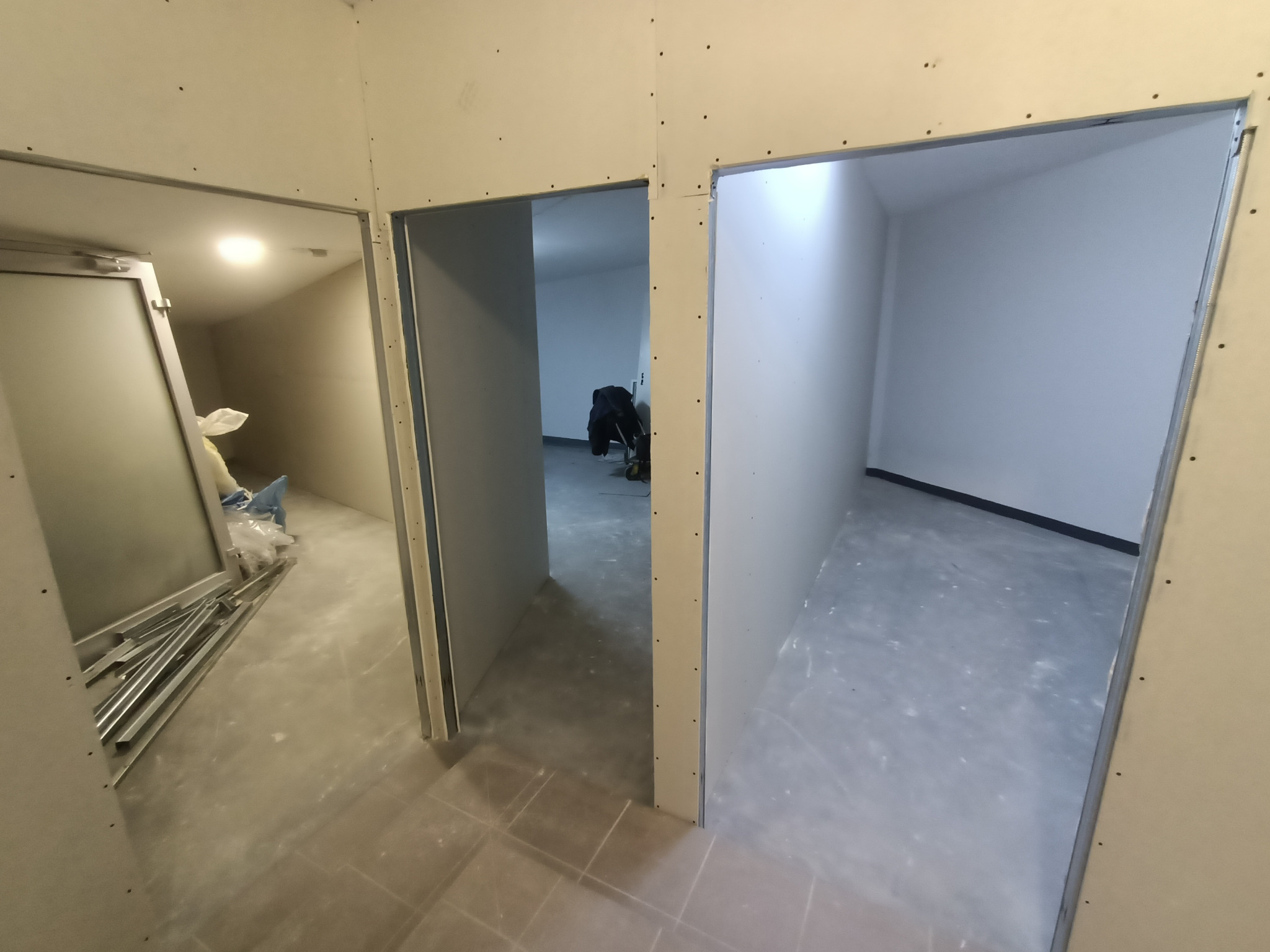 Wnętrze w trakcie prac wykończeniowych, widoczne surowe ściany z płyt gipsowo-kartonowych z metalowymi profilami, trzy otwory drzwiowe prowadzące do różnych pomieszczeń, na podłodze leżą materiały...