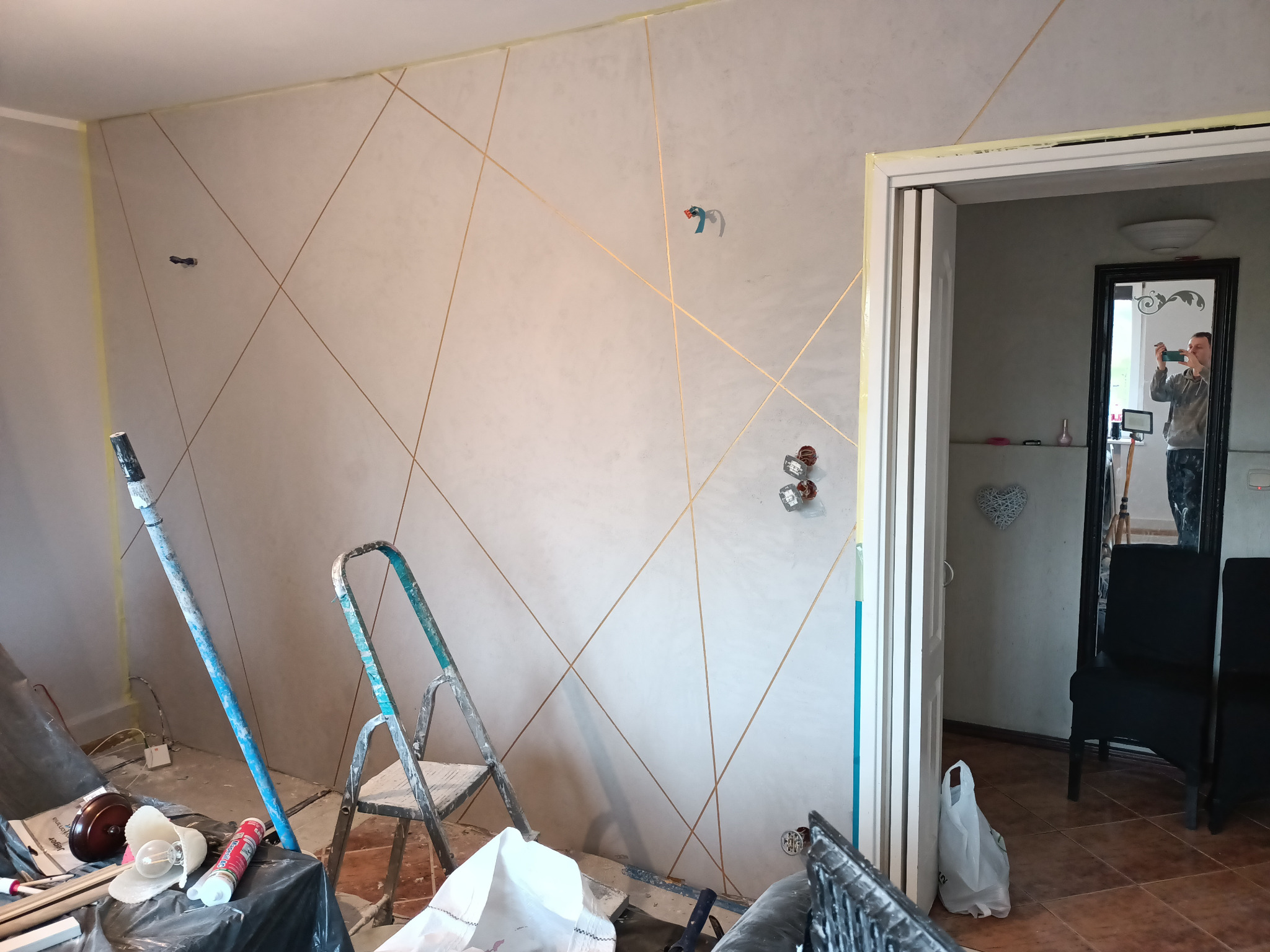 Ściana po szpachlowaniu z dekoracyjnymi złotymi liniami, widoczne ślady po taśmie malarskiej, drabina malarska i bałagan w trakcie prac wykończeniowych.