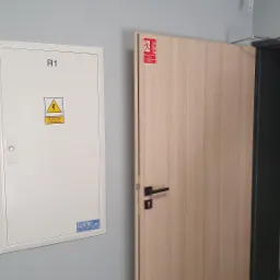 Biała skrzynka elektryczna z napisem 'R1' i ostrzeżeniem o napięciu, zamontowana na szarej ścianie obok drzwi z oznaczeniem ewakuacyjnym.