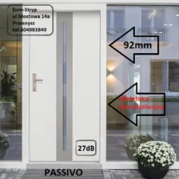 Białe drzwi wejściowe z szarymi panelami bocznymi i pionowym przeszkleniem, widoczne oznaczenia termoizolacyjności ościeżnicy, grubości 92mm oraz tłumienia dźwięku 27dB, przed drzwiami wycieraczka...