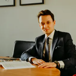 Młody mężczyzna w garniturze siedzi przy okrągłym drewnianym stole z otwartymi dokumentami i katalogiem, spoglądając w obiektyw.