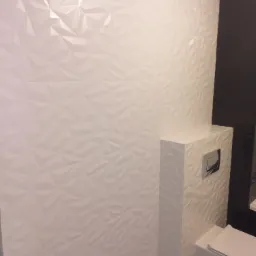 Nowoczesna łazienka z białą, trójwymiarową glazurą ścienną i podwieszaną toaletą z chromowanym przyciskiem spłukującym, kontrastująca z ciemną płytką po prawej stronie.