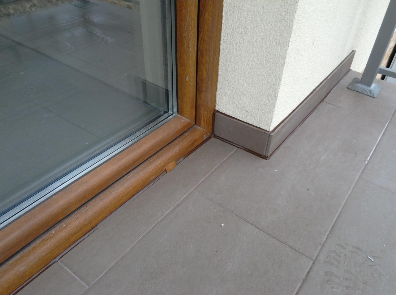 Szare płytki podłogowe na balkonie, brązowe cokoły przy ścianie i drewniana rama okna balkonowego.