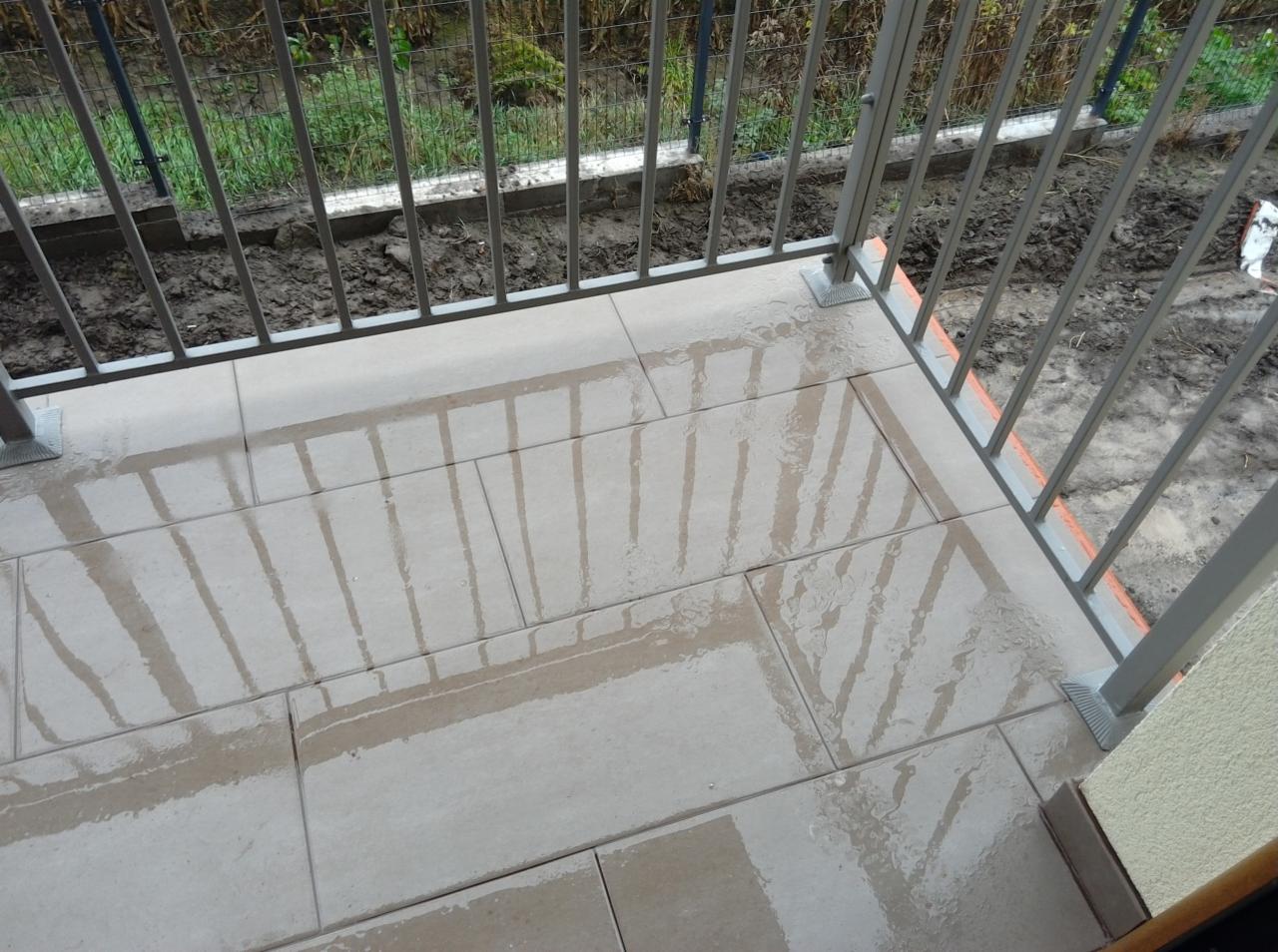 Świeżo ułożone, mokre płytki na balkonie z szarymi barierkami, odbijające się w wodzie, z widocznym fragmentem elewacji budynku.