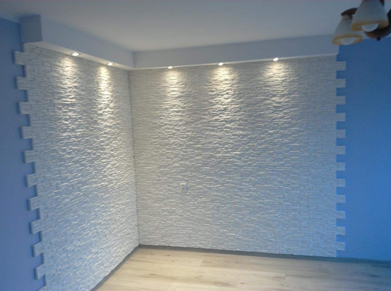 Ściana wykończona dekoracyjną białą okładziną imitującą kamień, z punktowym oświetleniem sufitowym, narożnik wykończony ozdobnymi płytkami w kształcie cegiełek.