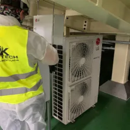 Pracownik w kombinezonie i żółtej kamizelce z logo GMK Tech instaluje jednostkę zewnętrzną klimatyzacji LG Multi V w budynku komercyjnym, widoczne elementy konstrukcyjne budynku.