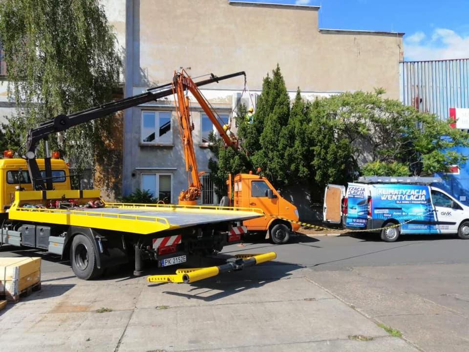 Dwa dźwigi samochodowe, żółty i pomarańczowy, podnoszą białą jednostkę zewnętrzną klimatyzacji przy budynku. W tle zaparkowany van z reklamą usług klimatyzacyjnych i wentylacyjnych.