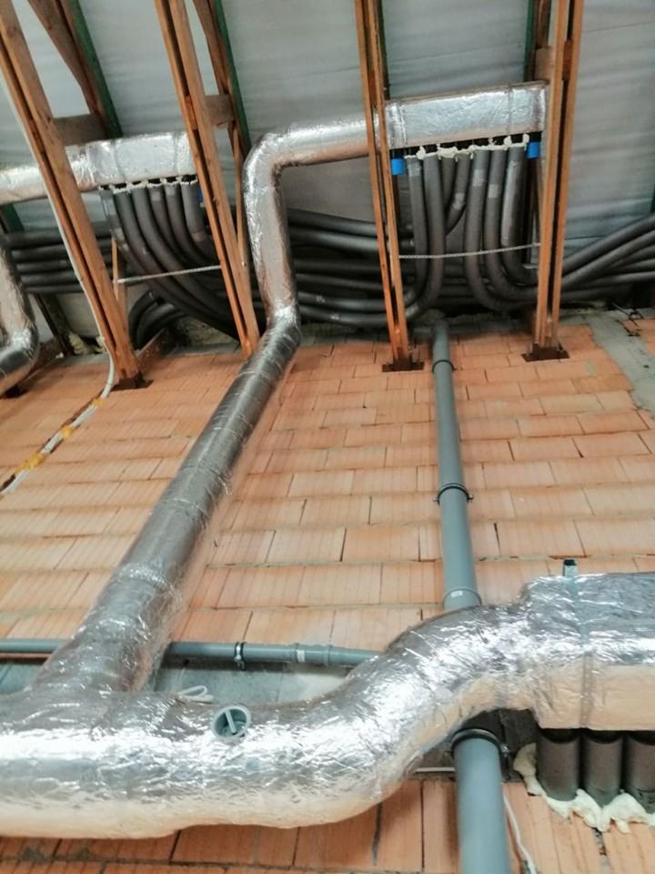 Instalacja rekuperacji w trakcie montażu na poddaszu, widoczne rury izolowane folią aluminiową, rozprowadzenie przewodów wentylacyjnych, ceglana podłoga i drewniana konstrukcja dachu.