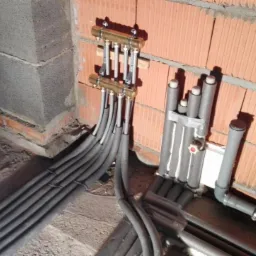 Rozdzielacz ogrzewania podłogowego z rurami w izolacji, zamontowany na ścianie z cegły dziurawki w surowym stanie, widoczne elementy instalacji hydraulicznej i wylewka podłogowa.