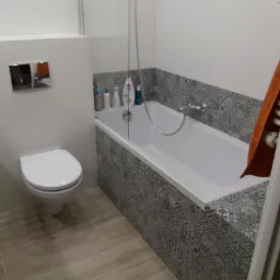 Nowoczesna łazienka z wanną obudowaną mozaiką w odcieniach szarości, wiszącą toaletą i podłogą z płytek imitujących drewno.