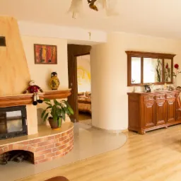 Salon z kominkiem obudowanym cegłą, drewnianą komodą z lustrami, stołem i krzesłami, z widokiem na sypialnię z żółtą dekoracją na ścianie.