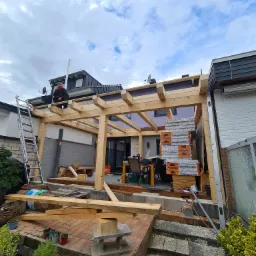 Konstrukcja drewnianej pergoli w trakcie budowy, widoczny mężczyzna siedzący na belce, stos materiałów budowlanych i narzędzia, na pierwszym planie rośliny w donicach.