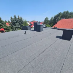 Nowoczesny dach pokryty szarą papą termozgrzewalną z widocznymi kominami wentylacyjnymi i pracującym dekarzem w czerwonej koszulce, na tle domów z czerwonymi dachami i zielonych drzew