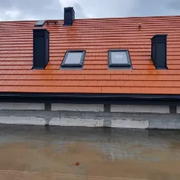 Nowy, pomarańczowy dach z czarnymi kominami i oknami dachowymi w Miłakowie, widziany z poziomu surowego stropu.