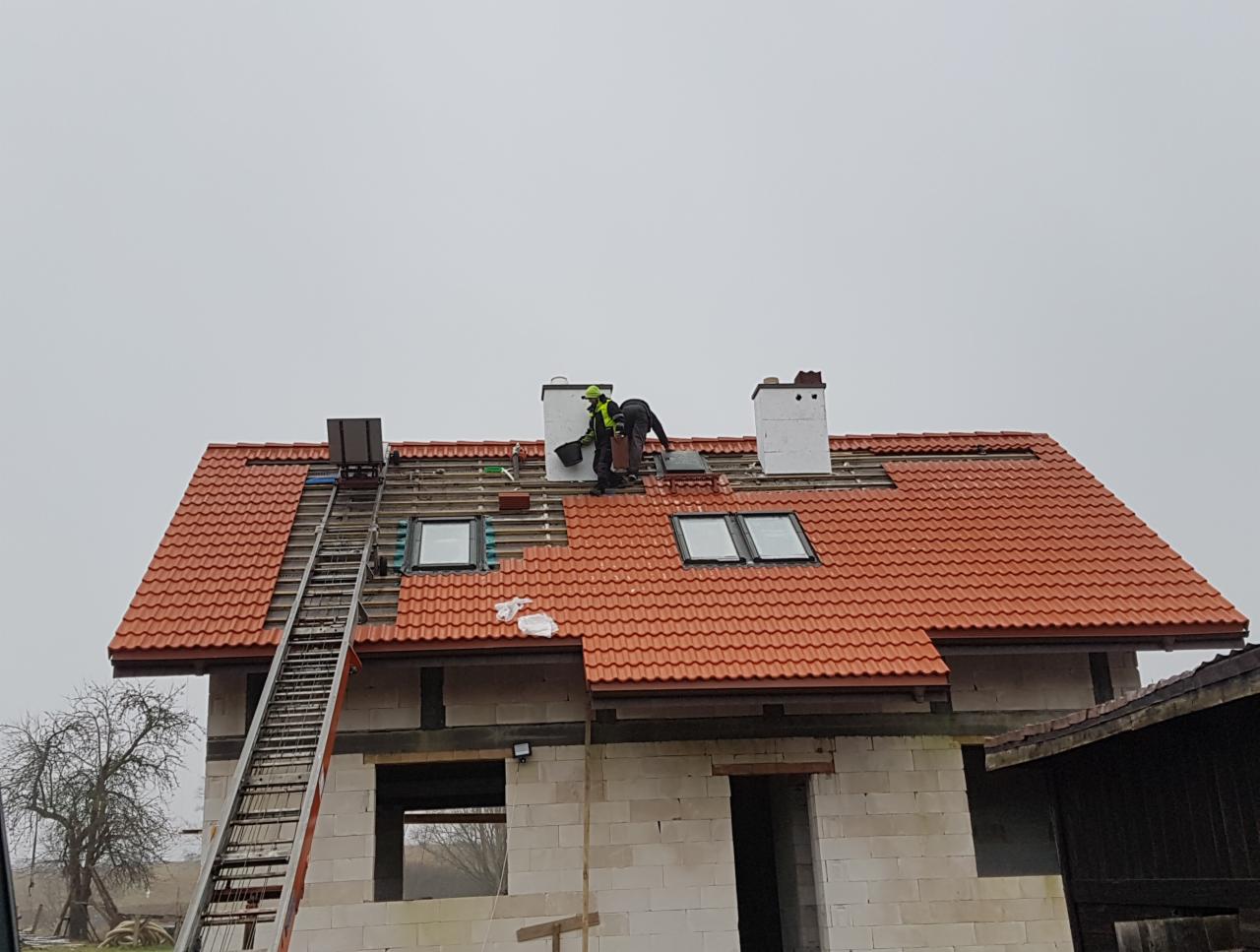 Montaż czerwonej dachówki na domu jednorodzinnym z oknami dachowymi, widoczna drabina oparta o dach oraz pracownicy w trakcie pracy przy kominie.