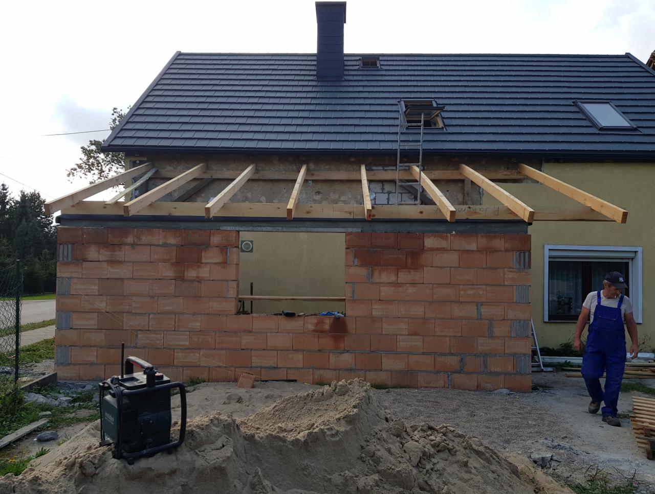 Budowa dachu: murowana konstrukcja z wystającymi krokwiami, widoczny fragment starego dachu i pracownik w kombinezonie na tle budynku.