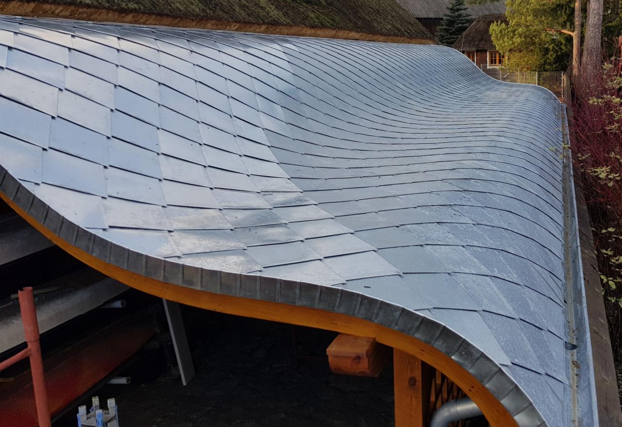 Falista konstrukcja z metalowych płytek dachowych, widok z góry na drewnianą konstrukcję nośną, w tle budynek kryty strzechą.