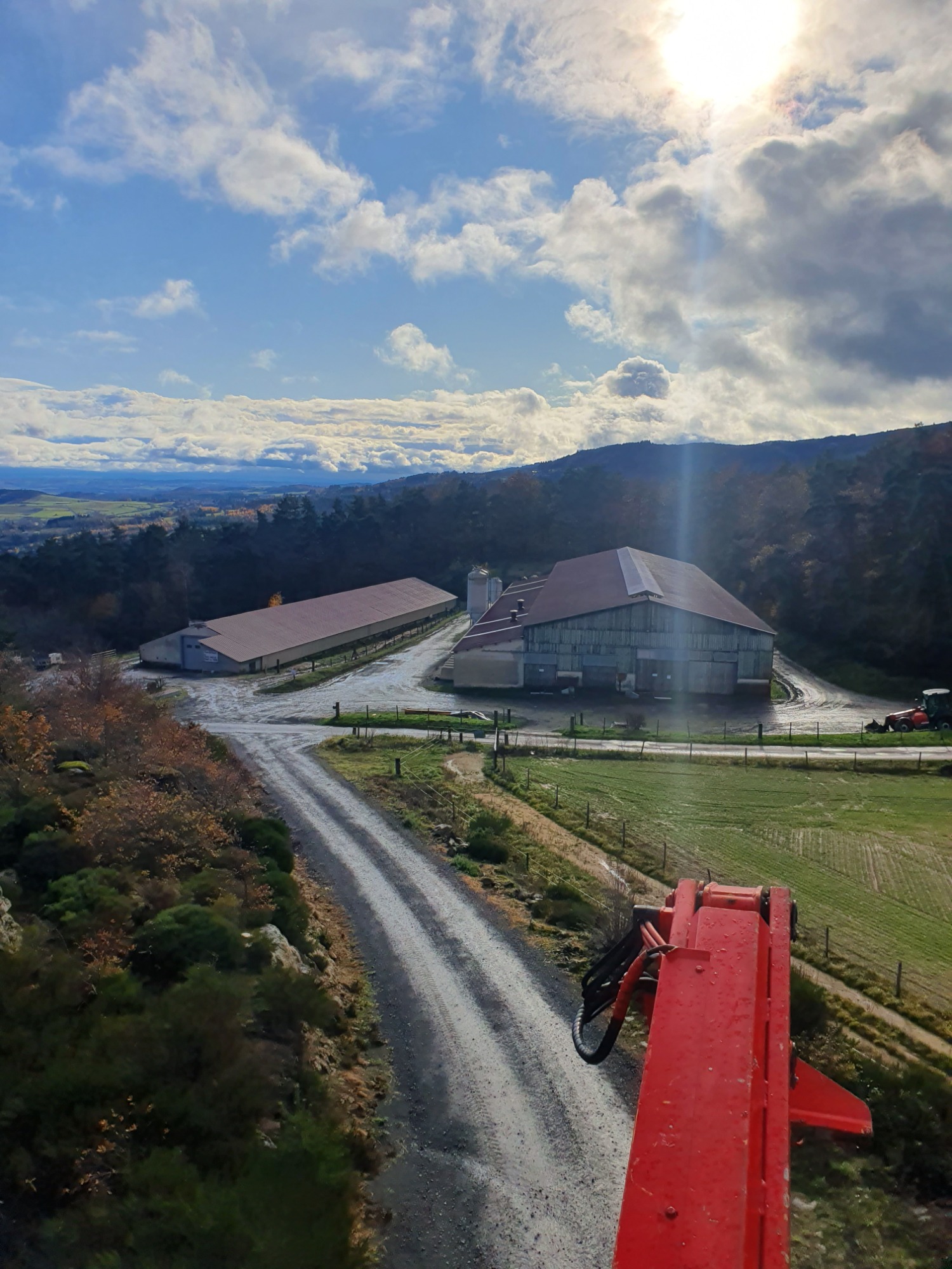 Widok z góry na zabudowania gospodarcze we Francji, z perspektywy podnośnika koszowego, w tle pagórkowaty krajobraz pod błękitnym niebem z chmurami.