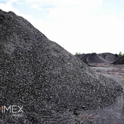 Góra ekogroszku z logo firmy POLIMEX w lewym dolnym rogu, na placu składowym z widocznymi śladami opon i innymi hałdami w tle.