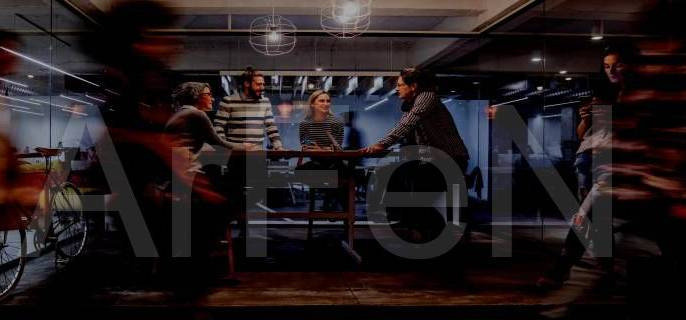 Zespół w nowoczesnym biurze, dyskutujący przy wysokim stole, z rowerem widocznym w tle za szklaną ścianą.