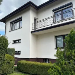 Jasna elewacja domu jednorodzinnego z balkonem, brązowe okna i dach, zadbany ogród z krzewami.