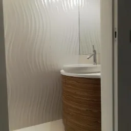 Nowoczesna łazienka z umywalką na szafce o zaokrąglonym kształcie i wzorze drewna, białe płytki ścienne z falistym wzorem, widok zza framugi drzwi.