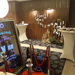 Fotobudka w lustrzanej ramie z wyświetlanym tekstem 'Co za wesele, miłość wisi w powietrzu' na tle dekoracji weselnej z lampionami, zdjęciami na sznurkach i rekwizytami na stoliku.