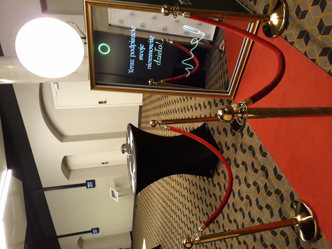 Interaktywne lustro z napisem 'Teraz podpiszcie swoje niesamowite dzieło!' otoczone czerwonymi słupkami z pozłacanymi elementami, ustawione na wzorzystej podłodze.