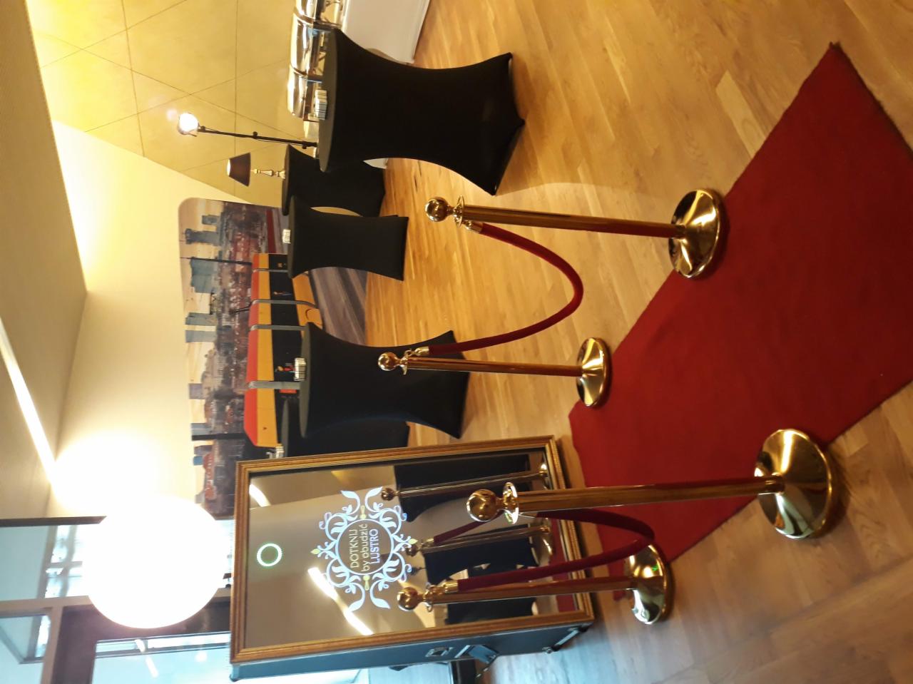 Fotobudka lustro z napisem 'Dotknij by obudzić lustro' otoczona złotymi słupkami z czerwonymi sznurami na czerwonym dywanie, w tle stoliki koktajlowe z czarnymi pokrowcami i panorama miasta.