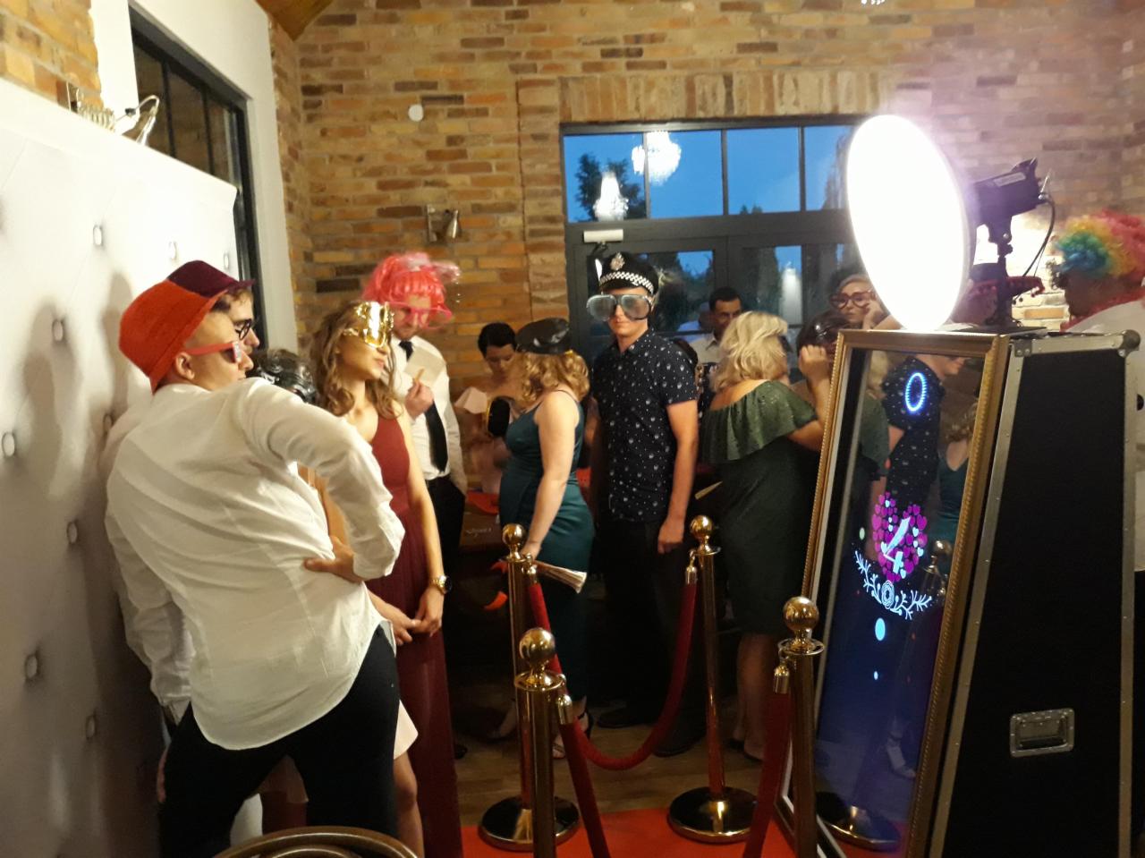 Grupa osób pozuje do zdjęcia wewnątrz pomieszczenia z ceglaną ścianą, korzystając z lustrzanej kabiny z rekwizytami, takimi jak peruki i okulary.